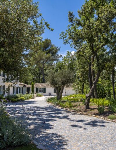 Allée pavée entourée d’arbres et de massifs fleuris menant à une maison provençale
