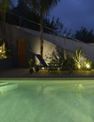 Illuminated pool at dusk with wooden deck, lounger and lit vegetation