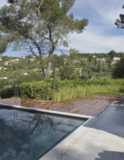 Piscine à débordement avec terrasse bois, chaises longues et jardin en arrière-plan