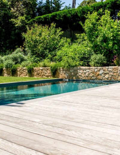 Pool bordered by a wooden deck with stone wall and flowering garden