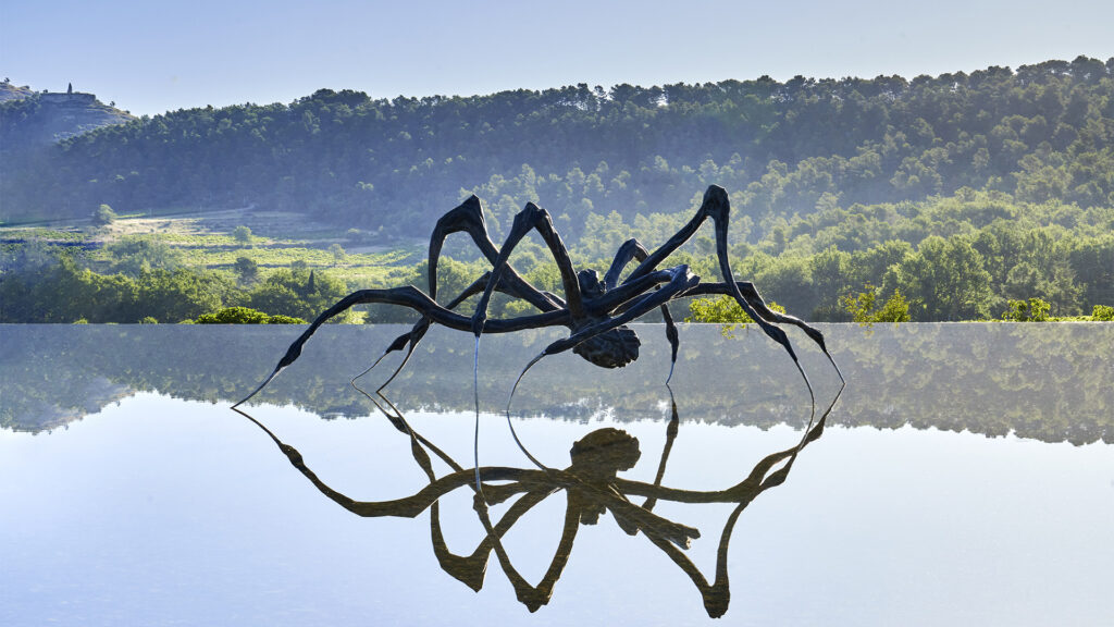 Metal spider sculpture, dramatically staged with a lush green landscape in the background