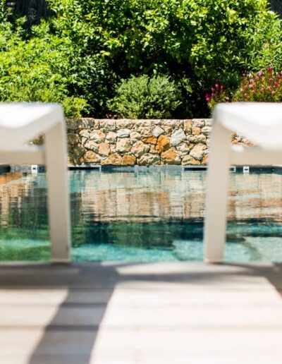 View of the pool from white sun loungers with a stone wall and garden in the background