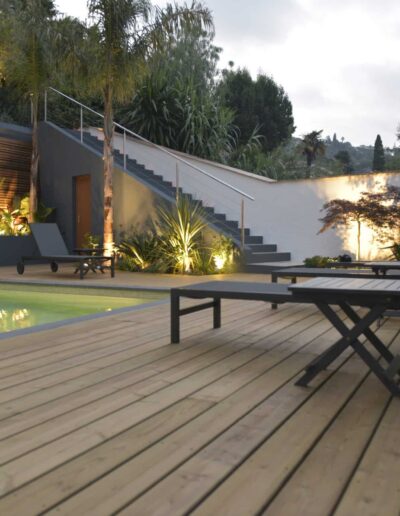 Wooden deck with illuminated pool, loungers and lit plants in the evening