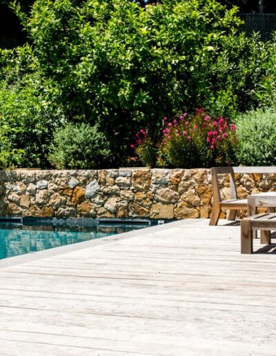 Wooden deck beside a pool with a stone wall and Mediterranean vegetation